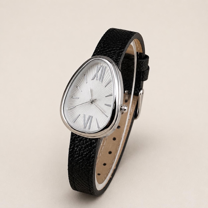 May include: A wristwatch with a teardrop-shaped silver case and a white sunburst dial featuring silver Roman numerals at the twelve and six o'clock positions. The watch has a textured black leather strap with a silver buckle and a light beige lining.