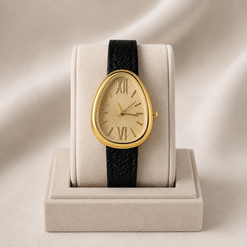May include: A wristwatch with a teardrop-shaped gold case and a textured gold dial featuring Roman numerals at the twelve and six o'clock positions. The watch has a black leather strap with a pebbled grain texture and a small gold crown on the side.