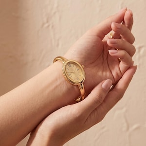 May include: A gold-toned bangle watch featuring an oval-shaped case with a smooth, polished finish. The watch face is gold with black Roman numeral hour markers and thin, matching gold-toned hands. The band is a solid, rigid cuff style.