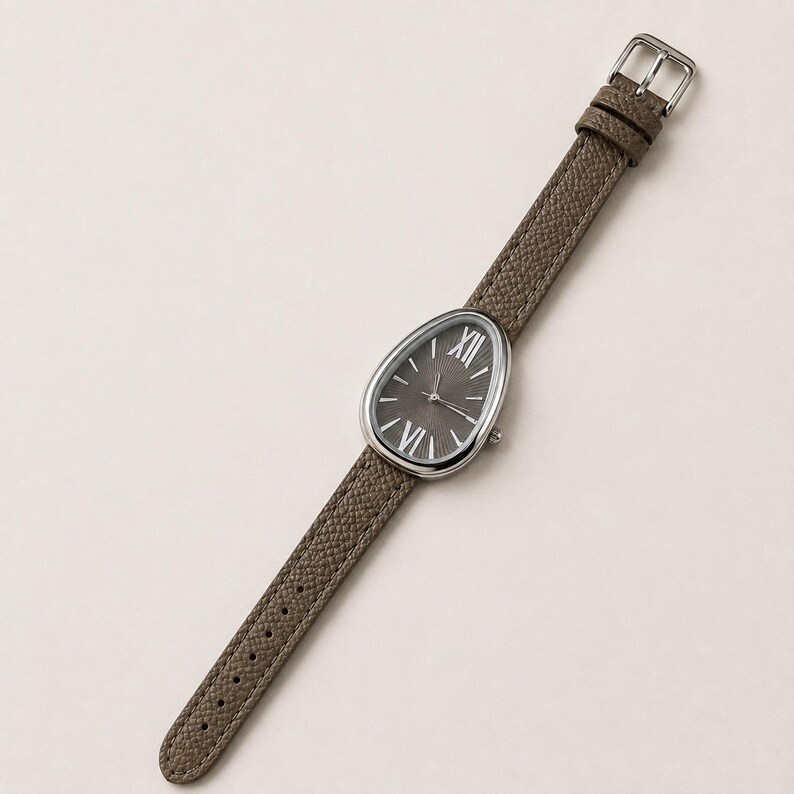May include: A wristwatch with a teardrop-shaped silver case and a dark gray dial featuring silver Roman numerals at the twelve and six o'clock positions. The watch has a textured taupe leather strap with a silver buckle and matching loops.
