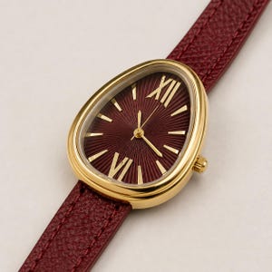 May include: A wristwatch with a gold-toned, teardrop-shaped case and a deep burgundy dial featuring a sunburst pattern, gold-toned Roman numeral indices, and thin gold hands. The watch is attached to a textured, dark red leather strap.
