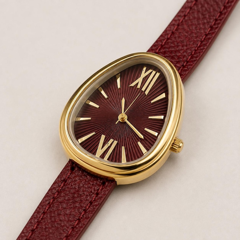 May include: A wristwatch with a gold-toned, teardrop-shaped case and a deep burgundy dial featuring a sunburst pattern, gold-toned Roman numeral indices, and thin gold hands. The watch is attached to a textured, dark red leather strap.