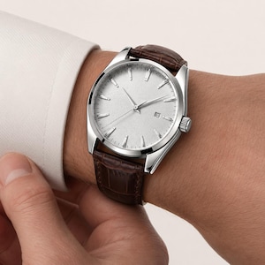 May include: A wristwatch featuring a silver-toned textured dial with slender hour markers, silver hands, and a date window at the three o'clock position. The watch has a polished silver-tone metal case and a dark brown leather strap with a crocodile pattern.
