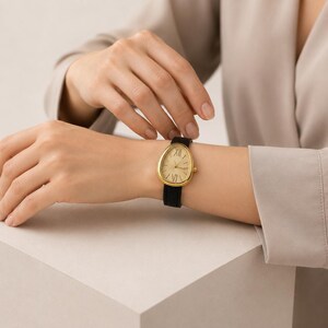 May include: A wristwatch with a teardrop-shaped gold-tone case and a textured champagne dial featuring Roman numerals at the twelve and six o'clock positions. The timepiece is secured with a slim black strap that has a subtle textured finish.