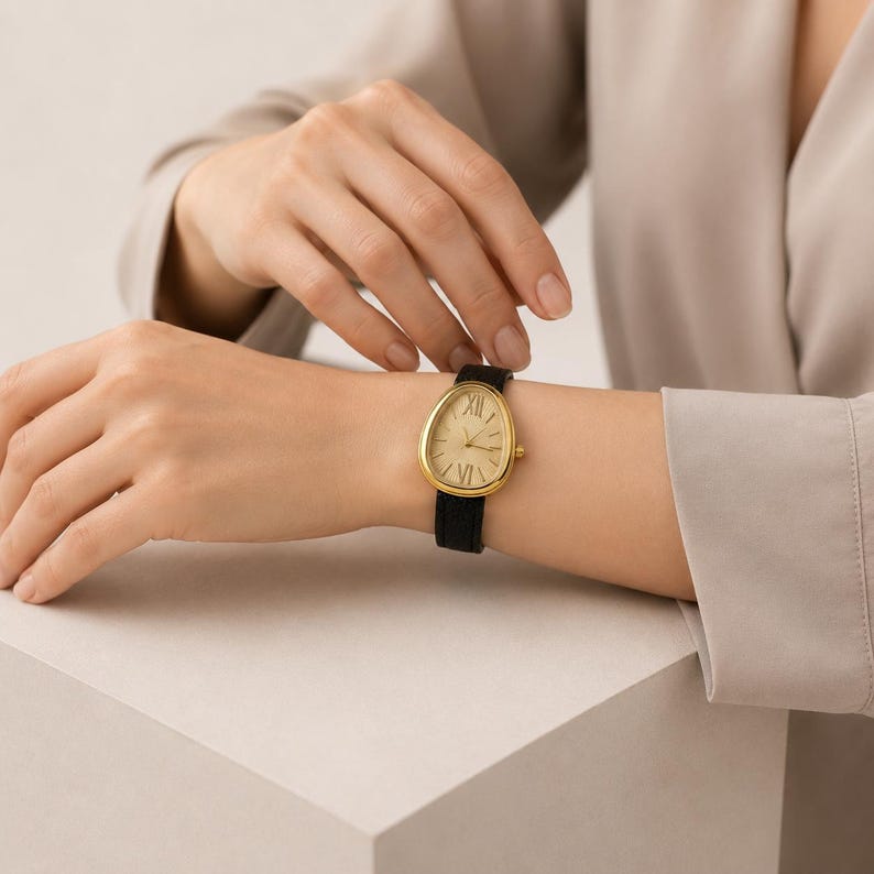 May include: A wristwatch with a teardrop-shaped gold-tone case and a textured champagne dial featuring Roman numerals at the twelve and six o'clock positions. The timepiece is secured with a slim black strap that has a subtle textured finish.