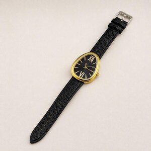 May include: A wristwatch with a teardrop-shaped gold-tone case and a black dial featuring gold-tone Roman numeral indices at the twelve and six o'clock positions. The watch includes a textured black leather strap with a silver-tone metal buckle.