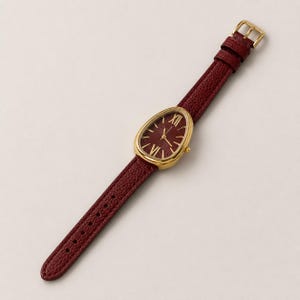 May include: A wristwatch with a teardrop-shaped gold-tone case and a dark red textured leather strap. The watch face is dark red with gold-tone Roman numeral hour markers at the twelve and six o'clock positions, and thin gold-tone hands.