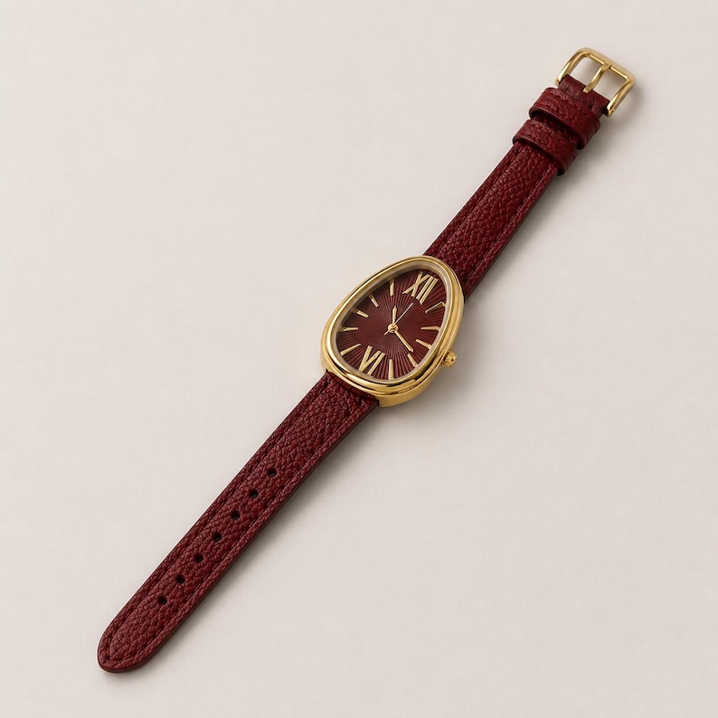 May include: A wristwatch with a teardrop-shaped gold-tone case and a dark red textured leather strap. The watch face is dark red with gold-tone Roman numeral hour markers at the twelve and six o'clock positions, and thin gold-tone hands.