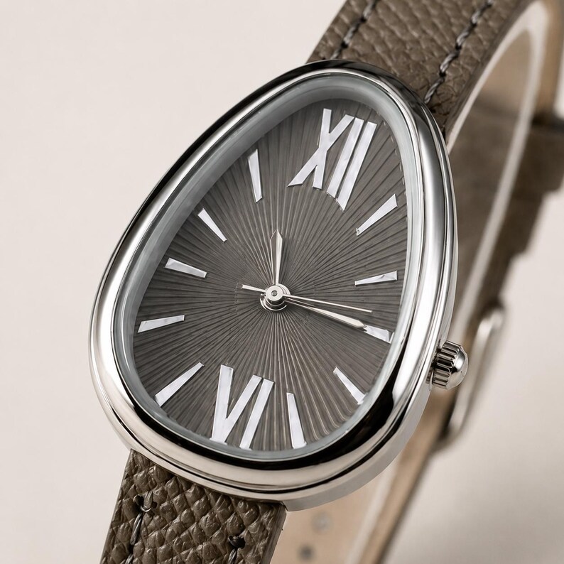 May include: A wristwatch with a teardrop-shaped silver case and a dark gray sunburst dial. The dial features white Roman numerals at the twelve and six o'clock positions, with simple silver hour markers. It has a textured taupe leather strap.
