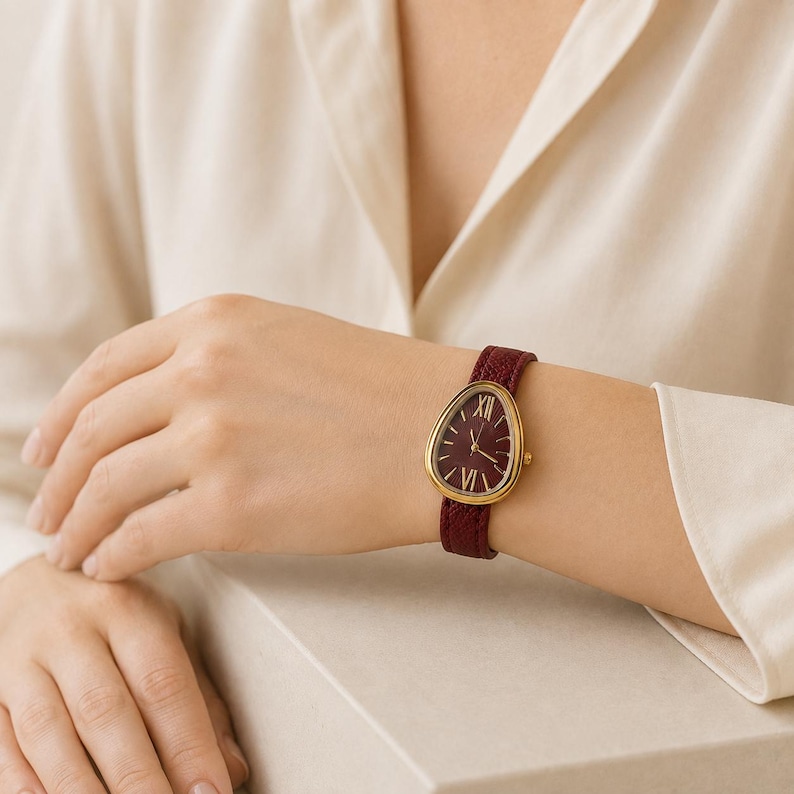 May include: A wristwatch featuring a teardrop-shaped gold case and a deep burgundy dial with gold Roman numeral markers at the twelve and six o'clock positions. The watch is finished with a textured burgundy leather strap with a gold buckle.