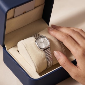 Könnte beinhalten: Eine silberfarbene Armbanduhr mit quadratischem Zifferblatt und flexiblem Metall-Dehnarmband. Die Uhr liegt auf einem cremefarbenen Samtkissen in einer dunkelblauen Präsentationsbox mit einer kleinen Karte im Deckel.
