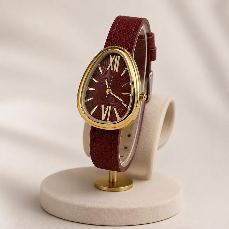 May include: A wristwatch with a teardrop-shaped gold-tone case and a deep red dial featuring gold Roman numeral indices at the twelve and six o'clock positions. The watch has a textured dark red leather strap with a gold-tone metal buckle.