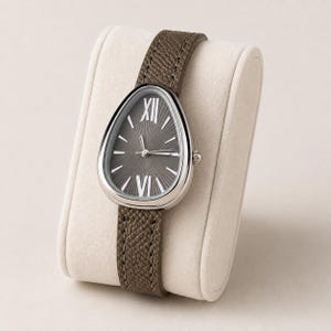 May include: A wristwatch with a teardrop-shaped silver case and a dark gray sunray dial featuring white Roman numerals at the twelve and six o'clock positions. The watch has a textured taupe leather strap with visible stitching along the edges.