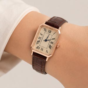 May include: A rectangular wristwatch with a rose gold-tone case and a cream-colored dial featuring black Roman numeral hour markers. The watch has dark blue hands and a brown leather strap with a textured, embossed crocodile-skin pattern.