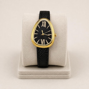 May include: A wristwatch featuring a teardrop-shaped gold-tone case and a black textured leather strap. The black dial displays gold-tone hour markers, including Roman numerals at the twelve and six o'clock positions, with gold-tone hands.