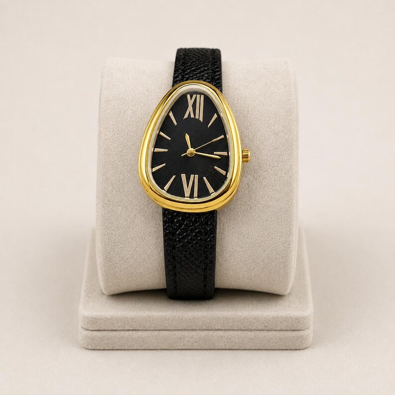 May include: A wristwatch featuring a teardrop-shaped gold-tone case and a black textured leather strap. The black dial displays gold-tone hour markers, including Roman numerals at the twelve and six o'clock positions, with gold-tone hands.
