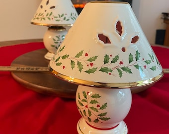May include: Two cream-colored porcelain candle lamps featuring a holly and berry pattern with gold-trimmed edges. The conical shades have cut-out leaf shapes, and the base is spherical. The Lenox brand name is printed on the shade.