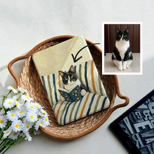 May include: A rectangular e-reader case featuring a painted illustration of a black and white tuxedo cat reading a blue book while sitting on a striped cushion. A small inset photo shows the real cat, and a bouquet of white daisies sits nearby.