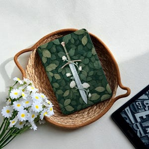 May include: A rectangular e-reader cover featuring a dark green background with a dense pattern of leaves and a central silver sword entwined with white roses. The cover is placed inside a round woven basket next to a bouquet of white daisies.