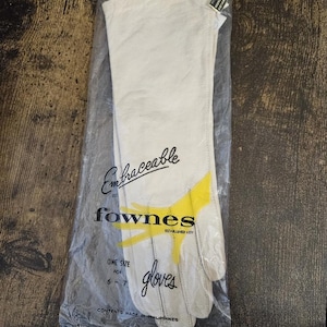 May include: A pair of off-white gloves inside a clear plastic bag. The packaging features the text Embraceable by fownes established 1777, one size for 6-7, and contents made in Philippines. A yellow hand graphic is printed on the front.