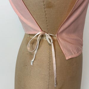 May include: A light pink top with a low back and a tie closure. The top is made of a lightweight fabric and is displayed on a mannequin. The tie is a thin, white ribbon.
