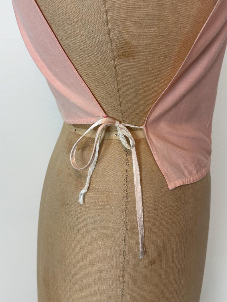 May include: A light pink top with a low back and a tie closure. The top is made of a lightweight fabric and is displayed on a mannequin. The tie is a thin, white ribbon.