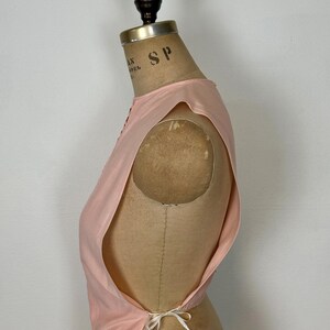 May include: A light pink sleeveless top with a delicate tie closure at the back. The top is displayed on a vintage dress form. The fabric appears to be lightweight, with a subtle sheen. The design features a scoop neckline and a tailored fit.