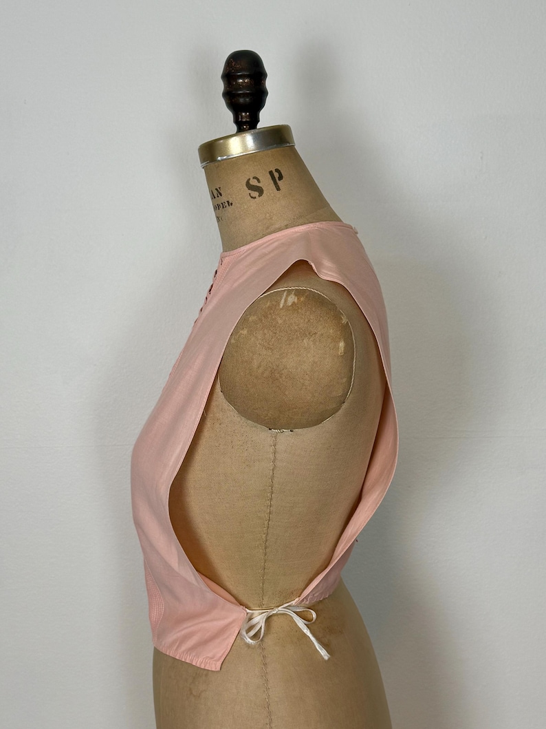 May include: A light pink sleeveless top with a delicate tie closure at the back. The top is displayed on a vintage dress form. The fabric appears to be lightweight, with a subtle sheen. The design features a scoop neckline and a tailored fit.