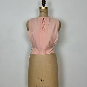 May include: A sleeveless, light pink top with a detailed, vertical lace-up design down the center. The top is displayed on a vintage dress form. White ties are visible on the sides. The dress form is tan and has the text "SP" and "Model 1966".