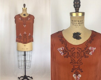 May include: A rust-colored vintage top with intricate beaded embellishments. The sleeveless blouse features a scoop neckline and a decorative design of dark beads and floral accents. The top is displayed on a vintage dress form.