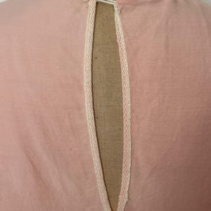 May include: Close-up of a light pink garment with a keyhole neckline. The neckline is trimmed with delicate cream-colored lace. A small, white button and loop closure is visible at the top of the opening. The fabric appears to be a soft, woven material.