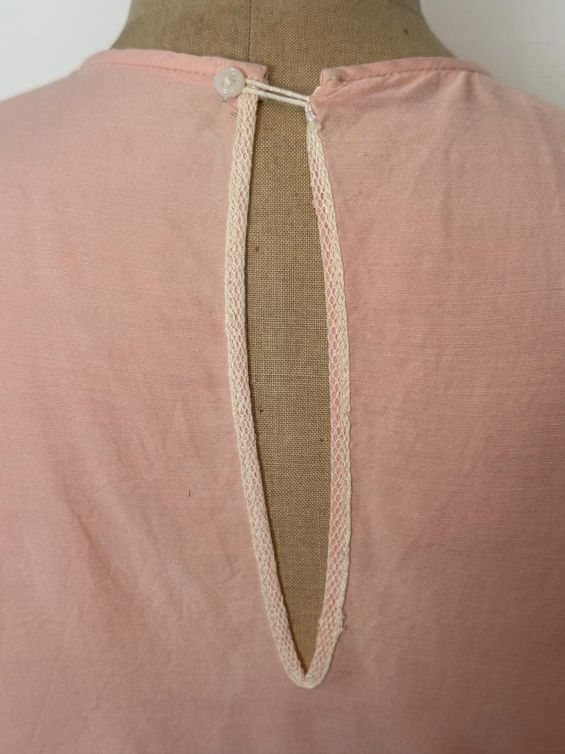 May include: Close-up of a light pink garment with a keyhole neckline. The neckline is trimmed with delicate cream-colored lace. A small, white button and loop closure is visible at the top of the opening. The fabric appears to be a soft, woven material.