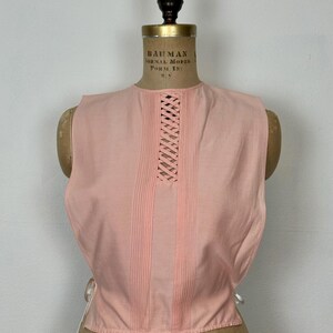 May include: A sleeveless, light pink top with a round neckline and a lattice design on the upper chest. Vertical pleats run down the front. White ties are visible on the sides. The top is displayed on a vintage dress form.