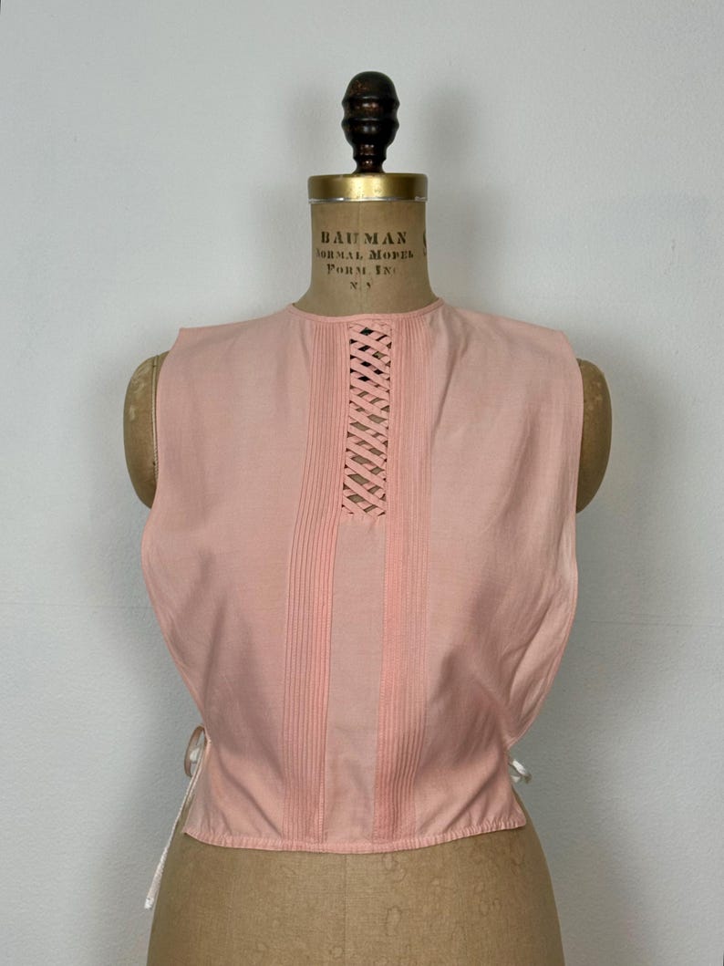 May include: A sleeveless, light pink top with a round neckline and a lattice design on the upper chest. Vertical pleats run down the front. White ties are visible on the sides. The top is displayed on a vintage dress form.