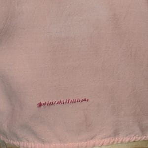May include: A light pink fabric with a soft, textured appearance. The fabric has a stitched border along the bottom edge and a small embroidered detail in a darker pink color.