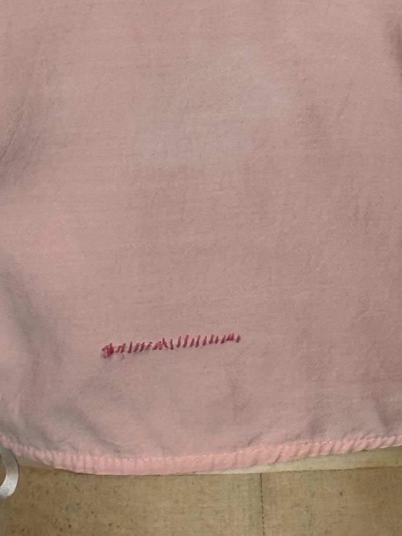 May include: A light pink fabric with a soft, textured appearance. The fabric has a stitched border along the bottom edge and a small embroidered detail in a darker pink color.