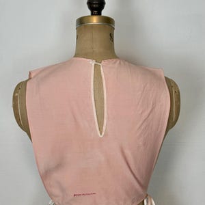May include: A light pink vintage top with a keyhole back and white trim. The top is displayed on a mannequin and has tie closures on the sides. Small red text is visible near the bottom.