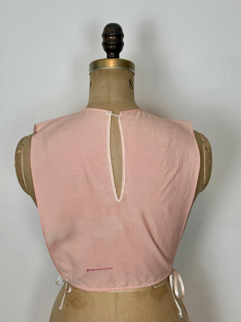 May include: A light pink vintage top with a keyhole back and white trim. The top is displayed on a mannequin and has tie closures on the sides. Small red text is visible near the bottom.