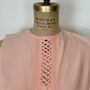 May include: A sleeveless, light pink top displayed on a vintage dress form. The top features vertical pleats on either side of a central, woven lattice design. The neckline is round, and the fabric appears to be a soft, lightweight material.