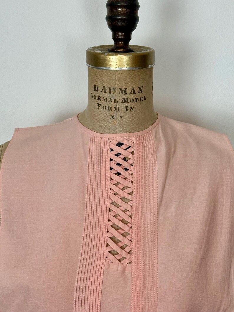 May include: A sleeveless, light pink top displayed on a vintage dress form. The top features vertical pleats on either side of a central, woven lattice design. The neckline is round, and the fabric appears to be a soft, lightweight material.