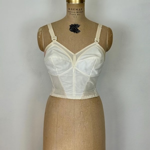 May include: A vintage white bustier with adjustable straps and a structured bodice. The bustier features a sheer panel at the neckline and is displayed on a vintage mannequin. The garment is a classic piece of lingerie.