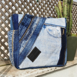 Puede incluir: Un bolso tote confeccionado con tela de mezclilla reutilizada en varios tonos de azul, con un diseño de patchwork con costuras diagonales, un bolsillo visible, bordes deshilachados y un parche rectangular negro de cuero sintético.