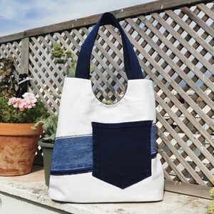 Puede incluir: Un bolso tote blanco con asas de mezclilla azul oscuro, una franja horizontal de mezclilla azul claro en la parte inferior y un bolsillo de parche de mezclilla azul oscuro centrado en el frente. El bolso tiene un diseño simple y abierto.