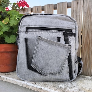 Puede incluir: Una mochila de mezclilla gris con lavado ácido, silueta rectangular, compartimento superior con cremallera horizontal y un bolsillo central grande. El diseño incluye detalles de tela negra desgastada en los bordes y las costuras.