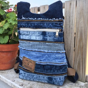 Puede incluir: Una mochila confeccionada con varios tonos de tela vaquera azul con bordes deshilachados. El bolso cuenta con un bolsillo horizontal con cremallera y un parche de cuero rectangular marrón con el texto ZARA MAN DENIMWEAR. Incluye correas de tela marrón.