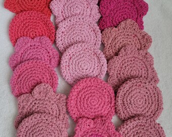 Puede incluir: Una colección de 17 posavasos de ganchillo en varios tonos de rosa y rojo. Los posavasos son todos redondos o con forma de flor y tienen una superficie texturizada.