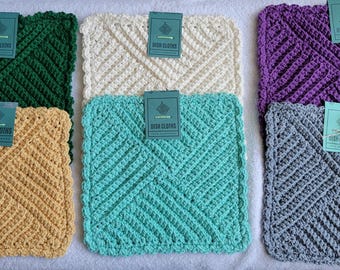 May include: A collection of six crocheted dish cloths in various colors: green, cream, purple, gray, yellow, and turquoise. Each cloth has a textured, geometric pattern and a tag that reads "Dish Cloths".