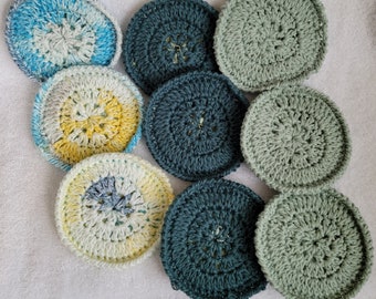 Op de afbeelding: Negen gehaakte onderzetters in verschillende kleuren en patronen. De onderzetters zijn rond en hebben een verhoogd midden. De kleuren omvatten blauw, groen, geel en wit.