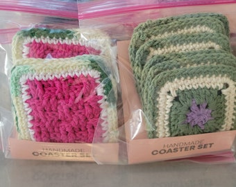 May include: Two sets of handmade crocheted coasters in plastic bags. One set is pink and green, the other is green and white with a purple flower in the center. Both sets have the text "Handmade Coaster Set" on the packaging.