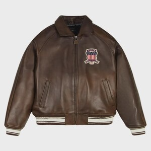 Puede incluir: Una chaqueta bomber de cuero marr&oacute;n con cierre de cremallera frontal, cuello en punta y dos bolsillos laterales. El pecho muestra un parche bordado con el texto AVIREX y una bandera estadounidense. Los pu&ntilde;os y el dobladillo tienen rayas blancas.